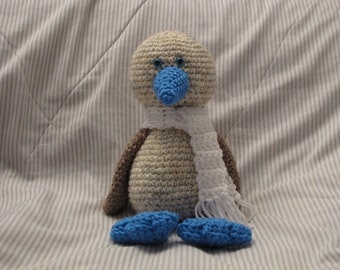 May include: A crocheted blue-footed booby bird toy with a white scarf. The bird is sitting on a striped fabric background.