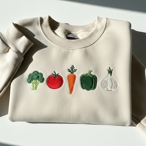 May include: A cream-colored sweatshirt featuring embroidered vegetables: broccoli, a tomato, a carrot, a bell pepper, and an onion. The vegetables are in green, red, orange, and white. The sweatshirt has a crew neck and ribbed cuffs.