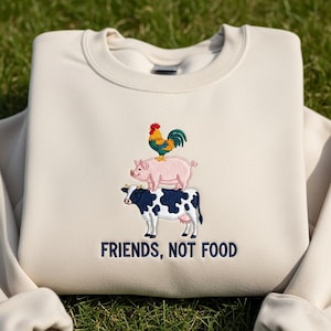 Op de afbeelding: Crèmekleurige sweatshirt met een geborduurd ontwerp van een koe, een varken en een haan op elkaar gestapeld. De tekst "FRIENDS, NOT FOOD" is eronder in marineblauw geborduurd.