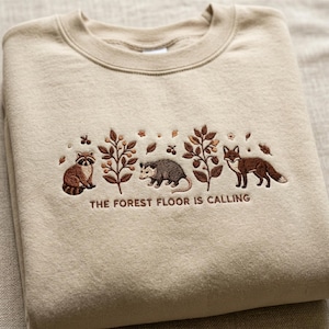 May include: Beige sweatshirt with embroidered forest scene. Features a raccoon, opossum, fox, and foliage. The text "THE FOREST FLOOR IS CALLING" is embroidered below the design. The sweatshirt is folded.