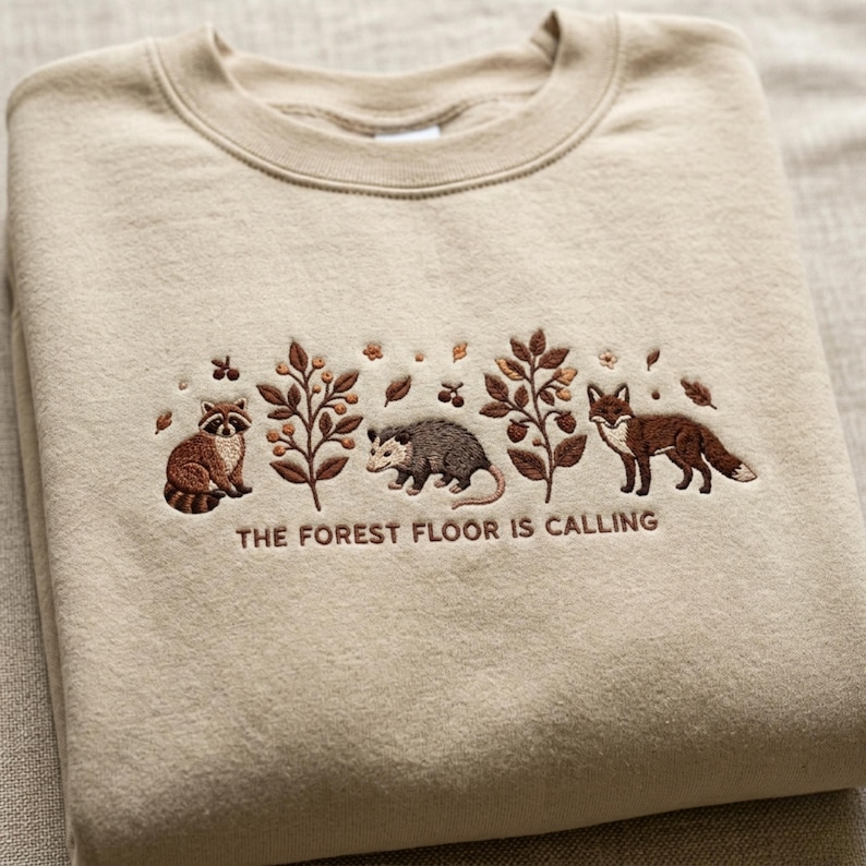 May include: Beige sweatshirt with embroidered forest scene. Features a raccoon, opossum, fox, and foliage. The text "THE FOREST FLOOR IS CALLING" is embroidered below the design. The sweatshirt is folded.