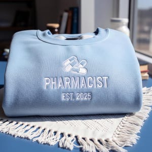 Op de afbeelding: Een lichtblauw sweatshirt met witte geborduurde tekst en pil-afbeeldingen. Het woord "PHARMACIST" wordt weergegeven, samen met "EST. 2025". Het sweatshirt is opgevouwen op een witte doek met franjes, met een blauwe achtergrond.