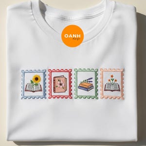 May include: White t-shirt with embroidered postage stamp designs featuring books and flowers. The stamps are in blue, red, green, and peach, with a sunflower, pink flowers, lavender, and orange flowers. The shirt has a crew neck.