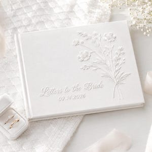 May include: A white rectangular book with an embossed floral design on the cover. The text "Letters to the Bride" and the date "09.14.2026" are embroidered in white thread. A small white ring box containing two gold rings is positioned nearby.