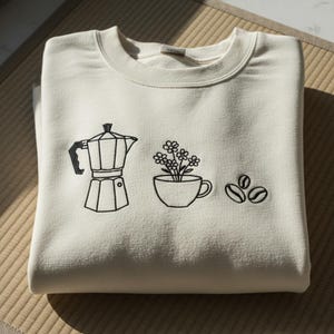 May include: A cream-coloured sweatshirt with a crew neck. The front displays a black line drawing of a coffee maker, a cup with flowers, and two coffee beans. The sweatshirt is folded on a woven mat.