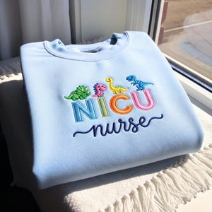 May include: A light blue sweatshirt with embroidered dinosaurs and the words "NICU nurse" in colorful letters. The design features a green, pink, yellow, and blue dinosaur above the text. The sweatshirt is folded on a white surface.