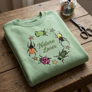 Op de afbeelding: Een lichtgroene sweatshirt met een geborduurd ontwerp met verschillende kleurrijke kevers en sterren in een cirkelvormige opstelling. De woorden "Nature Lover" zijn in een scriptlettertype binnen de cirkel geborduurd.
