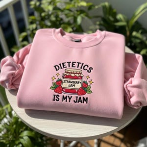 以下が含まれることがあります： 「DIETETICS IS MY JAM」の刺繍が入った薄いピンク色のスウェットシャツ。 イチゴジャムの瓶、イチゴ、星がデザインされています。 スウェットシャツは柔らかい素材で作られています。