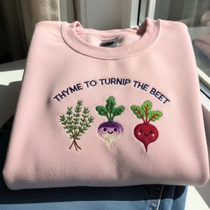 Peut inclure: Un sweat-shirt rose clair avec la phrase brodée "THYME TO TURNIP THE BEET". En dessous, des images brodées de thym, d'un navet violet et d'une betterave rouge, chacune avec des visages souriants.