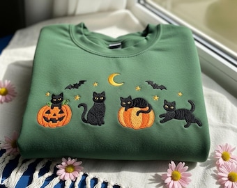 Puede incluir: Sudadera verde con diseño bordado de Halloween. Presenta gatos negros, calabazas, murciélagos, una luna creciente y estrellas. El diseño es en tonos naranja, amarillo y negro. La sudadera está doblada sobre una superficie blanca.