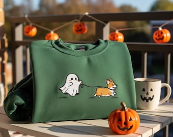 May include: A dark green sweatshirt featuring a cartoon ghost walking a corgi dog on a leash. A white mug with a jack-o'-lantern face and an orange pumpkin are on a white table. Halloween-themed decorations are in the background.