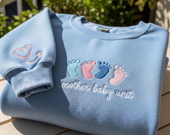 May include: Light blue sweatshirt with embroidered pink stethoscope on the sleeve. The front features embroidered baby footprints in shades of blue and pink, with the words "mother baby unit" in white script.