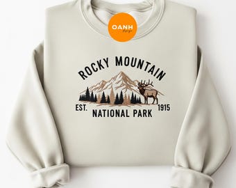 Può includere: Felpa color crema con la scritta "ROCKY MOUNTAIN NATIONAL PARK EST. 1915" in un design nero, marrone e verde. La grafica include una catena montuosa, pini e un cervo.