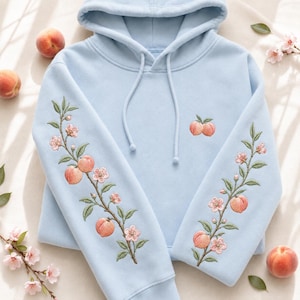 May include: A light blue hooded sweatshirt featuring detailed embroidery of peach branches with pink blossoms and orange peaches running down both sleeves, along with a small embroidered peach pair on the upper right chest area.
