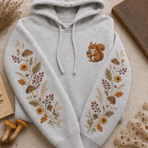 May include: A light grey hooded sweatshirt featuring detailed embroidery on both sleeves and the chest. The sleeves display botanical patterns of mushrooms, sunflowers, and berries. A small brown squirrel holding an acorn is embroidered on the chest.