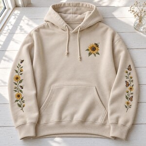 May include: A beige hooded sweatshirt featuring embroidered sunflower and bee designs on the left sleeve and chest, and a sunflower and butterfly pattern on the right sleeve. The garment includes a front kangaroo pocket and matching drawstrings.