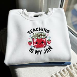 以下が含まれることがあります： 「TEACHING IS MY JAM」の刺繍が施された白いスウェットシャツ。デザインは、イチゴと青い星に囲まれた、緑と白の蓋が付いた赤いジャムの笑顔の瓶が特徴です。スウェットシャツは折りたたまれています。
