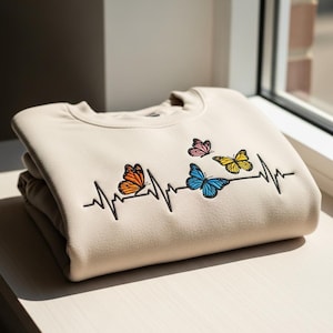 May include: Beige sweatshirt with a black heartbeat line design and colorful butterfly accents. The butterflies are orange, pink, blue, and yellow. The sweatshirt is folded and stacked on a light-colored surface.