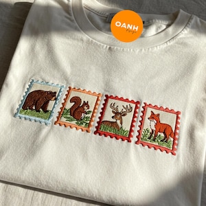 May include: White t-shirt featuring four embroidered postage stamp designs. Each stamp showcases a different woodland animal: a bear, squirrel, deer, and fox. The stamps have scalloped edges and are in various colors.
