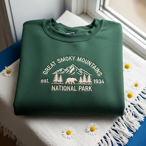 Könnte beinhalten: Ein dunkelgrünes Sweatshirt mit cremefarbener Stickerei "Great Smoky Mountains National Park". Das Design zeigt eine Bergkette, Bäume und einen Bären. Das Sweatshirt liegt gefaltet auf einem weißen Stoff mit Fransen.