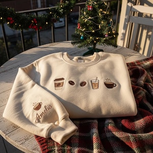 May include: Cream-colored sweatshirt with embroidered coffee-themed designs, including a to-go cup, coffee beans, and a steaming mug. The sleeve features a coffee cup and the name "Amelia" in script. The sweatshirt is displayed on a wooden table with a plaid blanket.