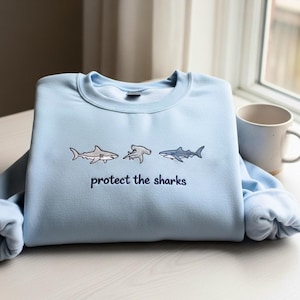 May include: Pale blue sweatshirt with embroidered shark designs. The sweatshirt displays three shark illustrations and the navy blue text "protect the sharks". A casual, comfortable top.