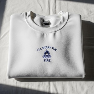 May include: White sweatshirt with a crew neck and the embroidered text "I'll Start the Fire" in navy blue. The design includes a bonfire graphic. The sweatshirt is folded neatly on a white surface.