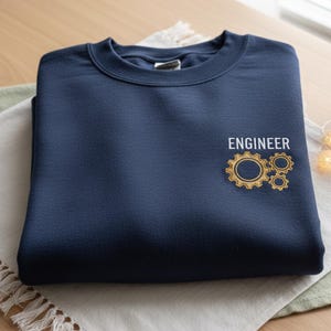 以下が含まれることがあります： 前面に「ENGINEER」の文字と金色の歯車デザインが刺繍されたネイビーブルーのスウェットシャツ。スウェットシャツは白と緑の布の上に折りたたまれています。