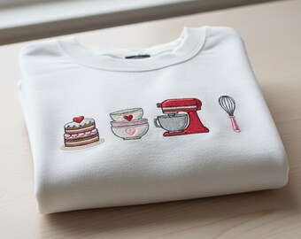 May include: White sweatshirt with embroidered baking-themed design. Features a cake, bowls, a stand mixer, and a whisk. The cake has a heart on top, and the bowls have heart details. The stand mixer is red, and the whisk is pink.