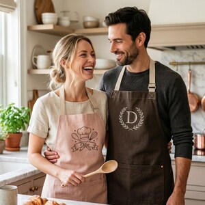 May include: A dusty pink apron featuring a chef hat, whisk, and spoon graphic with the name Chloe, and a dark brown apron featuring a letter D inside a laurel wreath. Both aprons have beige neck straps and waist ties, and front pockets.