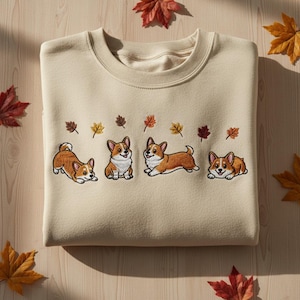 May include: Beige sweatshirt featuring embroidered corgi dogs and autumn leaves. The corgis are in various poses, with brown and white fur. The leaves are in shades of brown, yellow, and red. The sweatshirt is made of a soft material.