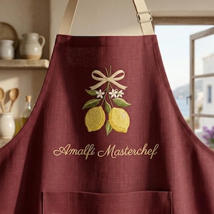 Peut inclure: Un tablier bordeaux profond orné d'une broderie représentant deux citrons jaunes avec des feuilles vertes et des fleurs blanches, noués par un ruban crème. Le texte "Amalfi Masterchef" est brodé en lettres dorées sous le motif de citrons.
