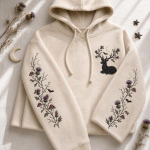 May include: A cream-colored hooded sweatshirt featuring intricate embroidery. The chest displays a black deer with antlers adorned with thistles, a crescent moon, and a bat. Both sleeves feature long, vertical botanical designs of thistles, mushrooms, and bats.