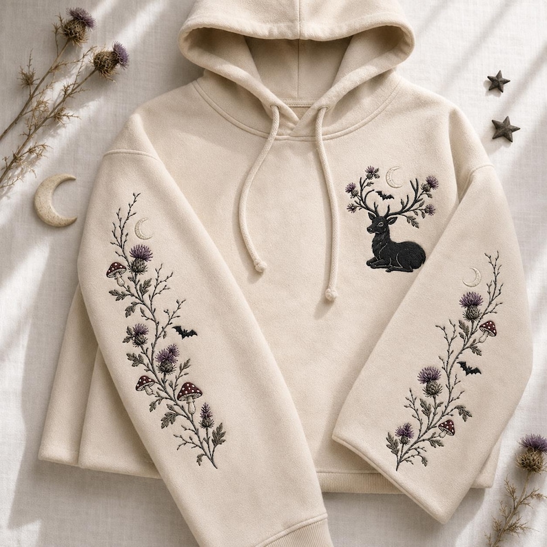 May include: A cream-colored hooded sweatshirt featuring intricate embroidery. The chest displays a black deer with antlers adorned with thistles, a crescent moon, and a bat. Both sleeves feature long, vertical botanical designs of thistles, mushrooms, and bats.