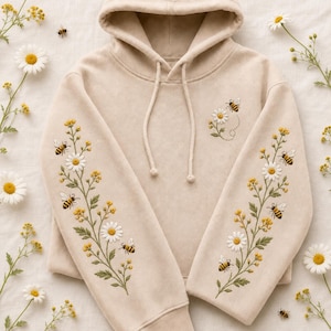 May include: A beige hooded sweatshirt featuring intricate embroidery of white daisy flowers, yellow wildflowers, and small honeybees along both sleeves and on the upper left chest. The hoodie includes matching beige drawstrings with rounded ends.