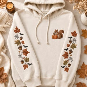 May include: A cream-colored hooded sweatshirt featuring embroidered designs. The chest displays a squirrel holding a pumpkin. Both sleeves are decorated with a vertical pattern of autumn leaves, acorns, spiderwebs, and small black bats.