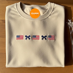 Könnte beinhalten: Cremefarbenes Sweatshirt mit gestickten amerikanischen Flaggen und einer blauen Schleife. Das Design zeigt zwei Flaggen, die eine Schleife in der Mitte flankieren. Das Sweatshirt hat einen Rundhalsausschnitt und lange Ärmel.