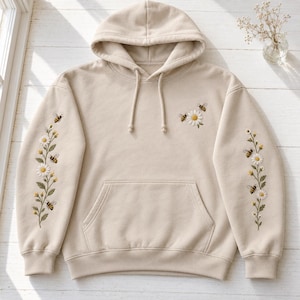 Peut inclure: Un sweat à capuche beige orné de motifs floraux et d'insectes brodés. Des marguerites blanches aux tiges vertes et de petites abeilles jaunes et noires sont cousues le long des manches et sur la poitrine gauche. Il possède une poche ventrale.