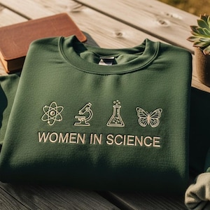 Pode incluir: Um moletom verde escuro com a frase bordada "WOMEN IN SCIENCE" e gráficos com tema científico. O design inclui um átomo, um microscópio, um béquer e uma borboleta. Um livro marrom e uma suculenta em vaso estão no fundo.