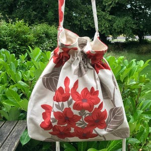 May include: A white drawstring bag with a red floral pattern. The bag has a long strap and is made of a soft, canvas-like material.