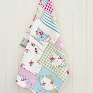 May include: A patchwork tea towel with a green, blue, pink, and white colour scheme. It features a teapot, teacups, and the text "two for tea" and "tea time".