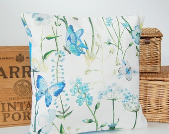 May include: A square scatter cushion featuring a white background with a botanical print of blue butterflies, green stems, and light blue flowers. The design includes delicate watercolour-style illustrations of various plants and insects scattered across the fabric.
