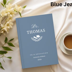May include: A blue jean-colored book with the text "Dr. Thomas" and a graduation cap and diploma graphic. The book also includes "PH.D. EDUCATION", "University of Miami", and the year "2026". A cup of coffee and flowers are in the background.
