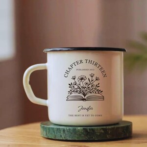 May include: White enamel mug with a black rim and handle. The mug features the text "CHAPTER THIRTEEN PUBLISHED 2013" in a semi-circle, with a floral design over an open book. The name "Jenifer" and the phrase "THE BEST IS YET TO COME" are also printed on the mug.