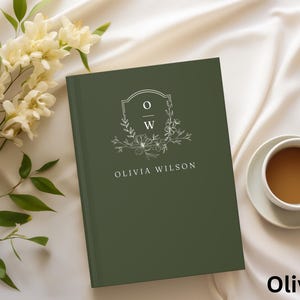 May include: An olive green journal with the name "Olivia Wilson" printed in white. The journal features a floral crest with the initials "OW". A cup of coffee and flowers are in the background.