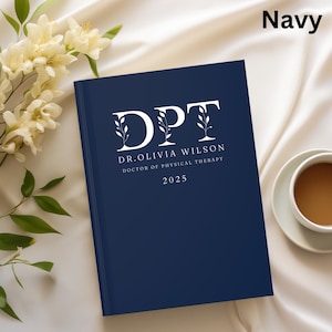 May include: A navy blue hardcover journal with white text that reads "DPT DR. OLIVIA WILSON DOCTOR OF PHYSICAL THERAPY 2025". The journal is next to a cup of coffee and a bouquet of white flowers.