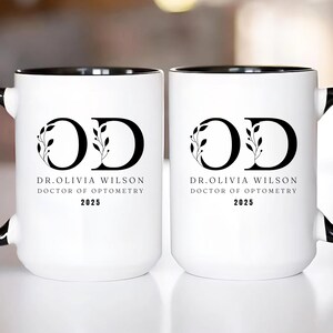 May include: Two white ceramic mugs with black handles and rims. Each mug features the letters "OD" in a black, stylized font, with botanical accents. Below the letters is the text "DR. OLIVIA WILSON DOCTOR OF OPTOMETRY 2025".