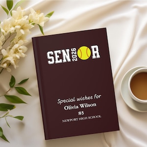 May include: A maroon notebook with the word "SENIOR" in white block letters and a yellow softball graphic. The text "2026" is above the softball. Below, it reads "Special wishes for Olivia Wilson #5 Newport High School."