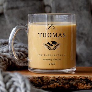 May include: A clear glass mug filled with a light brown beverage. The mug has a handle and features black text that reads "Dr. THOMAS" above a graduation-themed emblem and "P.H.D. EDUCATION, University of Miami, 2025."