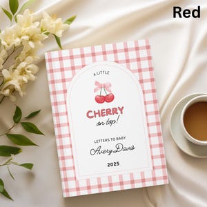 May include: A baby memory book with a red and white gingham pattern. The cover features the text "A Little Cherry on Top!" with a cherry illustration. The book includes the text "Letters to Baby", a name, and the year 2025.